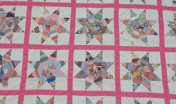 Detail of a bed quilt top made from a series of squares with a central mofit of eight-pointed stars. Each point of a star is made up of numerous small pieces of cloth. The stars are separated from each other by lines in plain pink cloth. USA, mid-20th century (TRC 2017.3349).