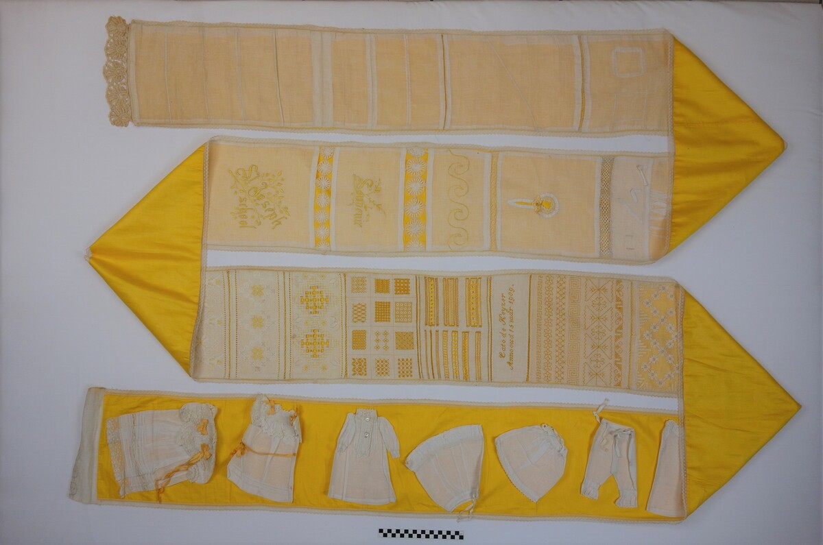 Sampler ('Pronkstuk') made in 1909 by Cato de Keijzer (TRC 2014.0938). The sampler is more than seven metres long. Below, and as part of the sampler, is a row of miniature garments.
