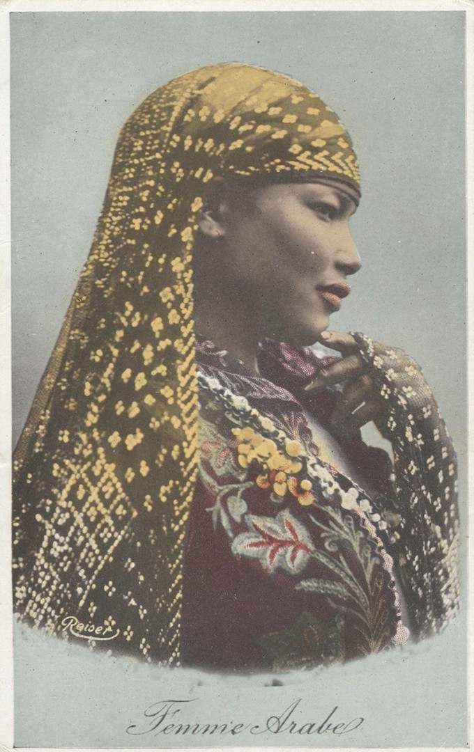 Early 20th century postcard from Egypt with a woman wearing an embroidered dress with necklaces, and a large black mantle decorated with metallic knots ('tulle-el-telle', 'badla') (TRC 2017.1844).