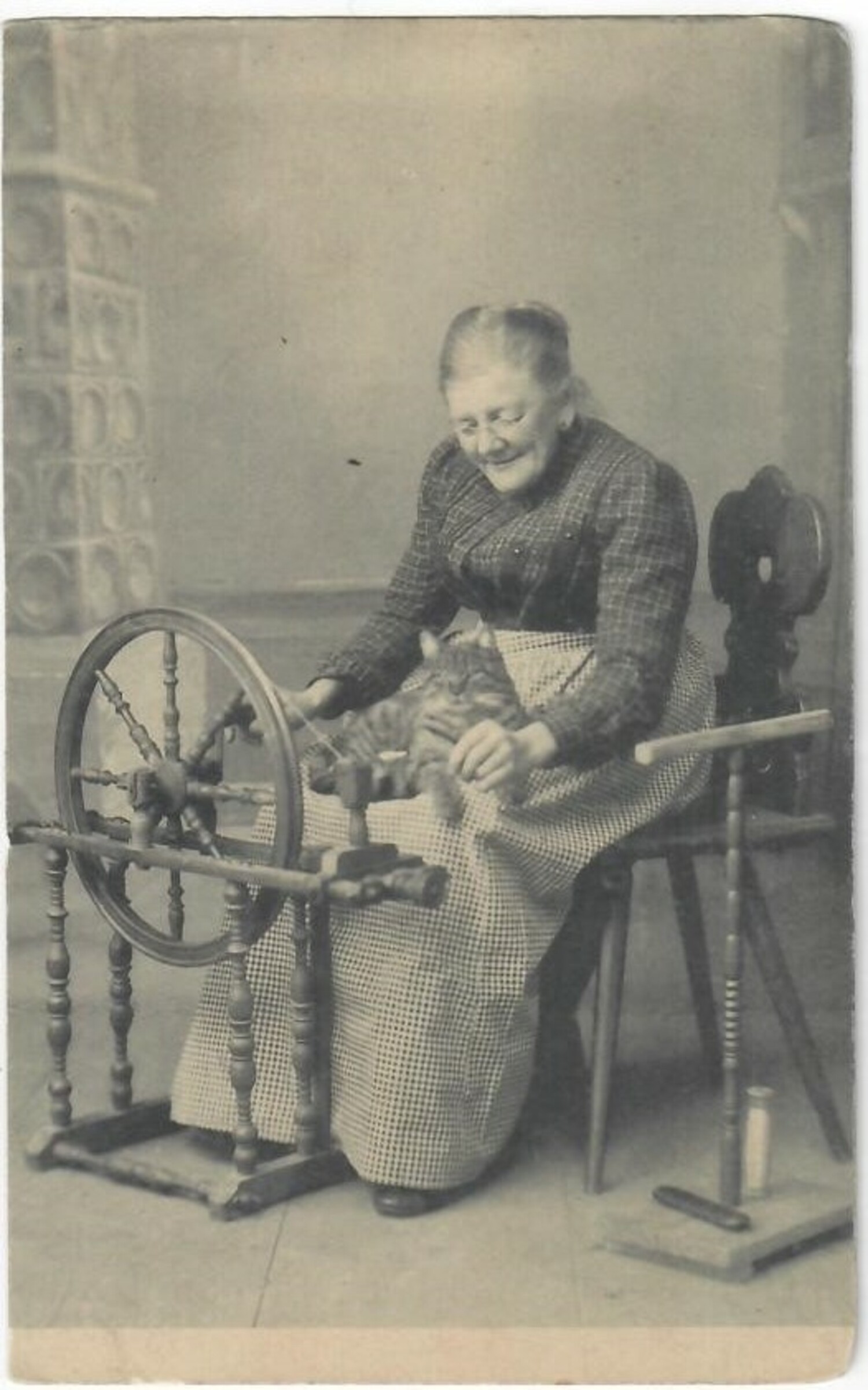 Early 20th century postcard showing an elderly woman with a cat on her lap sitting next to a horizontal spinning wheel. There is a large niddy-noddy on the floor beside her (TRC 2020.4314).