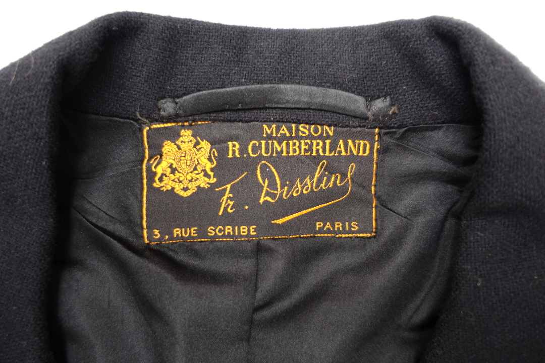 Label attached to long, black jacket with a deep split in the back. It is fastened at the waist with a single black button. France, 1935 (TRC  2021.3360a).