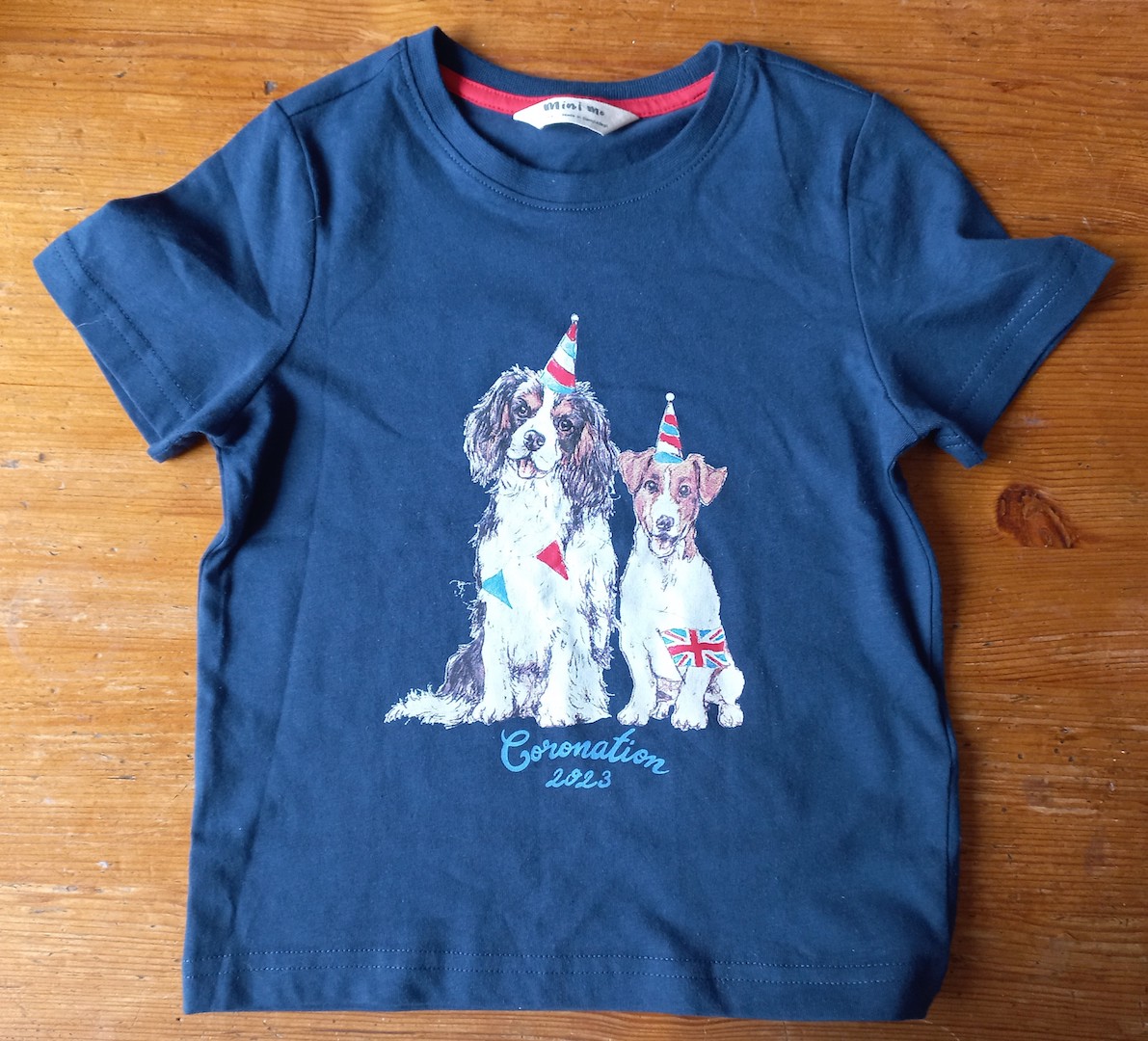 Small dark blue T-shirt for the 2023 Coronation, with a printed image of a King Charles spaniel and a Jack Russell terrier, both of which are wearing party hats, flags, etc. (TRC 2023.0679a).