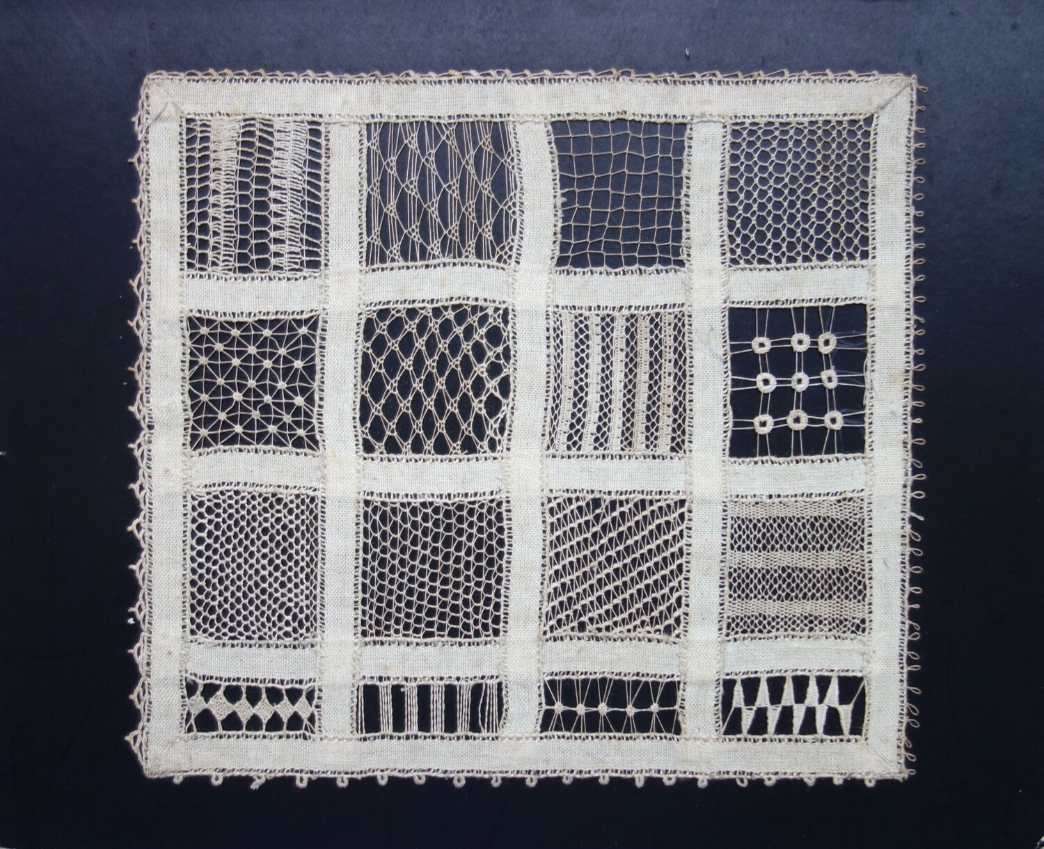 Piece of black card with a sample of a white frame made from tape lace that has been used to make 16 panels, each of which is filled with a different form of needle lace and net. Made by G.H.P. Wening betweeen 1919 and 1922 (TRC 2023.2805.094).