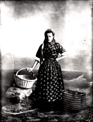 A studio portrait of a Cullercoats Fishwife in 1897. Kate Donkin has been photographed wearing her 'best' clothes which include a printed silk or cotton blouse with matching apron, a silk square filling the neckline of her blouse and a bonnet with the ribbons hanging loose. From Newcastle Libraries.