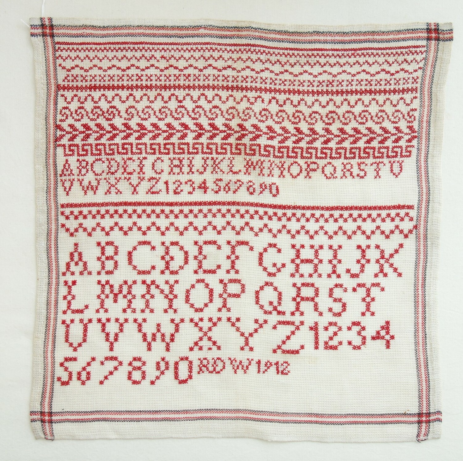 A school sampler worked with cross stitch and eyelet stitch using a red cotton yarn. The Netherlands, 1912 (TRC 2020.1609).